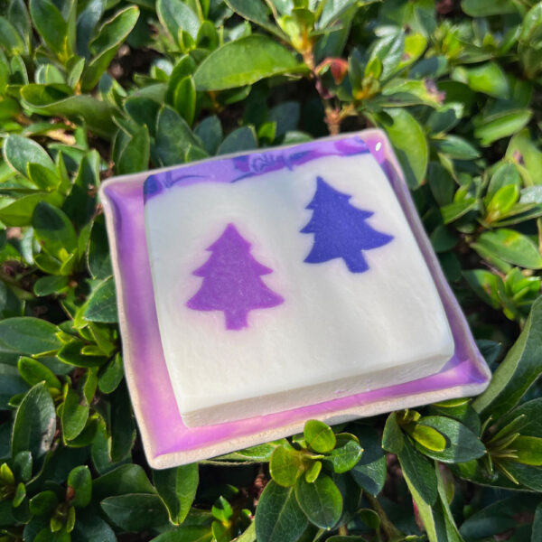 A lavender Christmas soap displayed on a square purple ceramic soap dish, with a background of dark green foliage