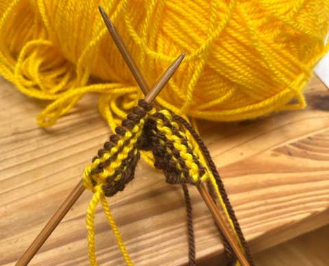 A ball of yellow yarn behind several rows of brown and yellow knitting on bamboo needless