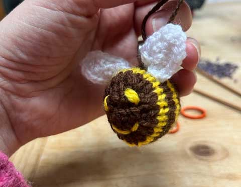 A knitted bee, stuffed with lavender, being held up by a hand and an out of focus wooden tabletop in the background