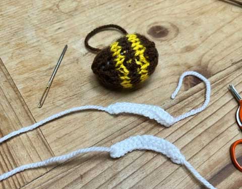 a knitted, stuffed bee body, with a wool needle and two knitted wings, ready to be stitched up and attached to the bee, set out on a wooden table top