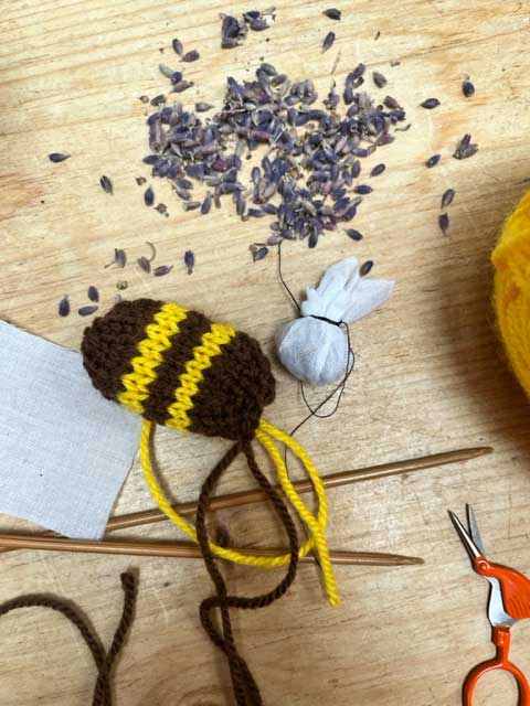 A wooden tabletop, with a scattering of lavender buds, a pair of bamboo needles, a made-up lavender sachet, a pair of orange sewing scissors with blades shaped like a bird's face, and some strands of yellow and brown yarn