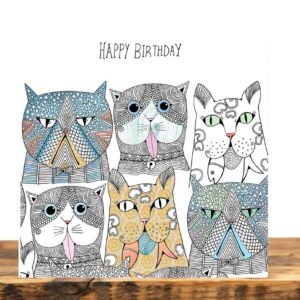 Greeting card on a wooden table. The card shows six intricately illustrated abstract cat faces with different expressions, below the words Happy Birthday.