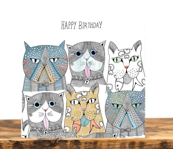 Greeting card on a wooden table. The card shows six intricately illustrated abstract cat faces with different expressions, below the words Happy Birthday.