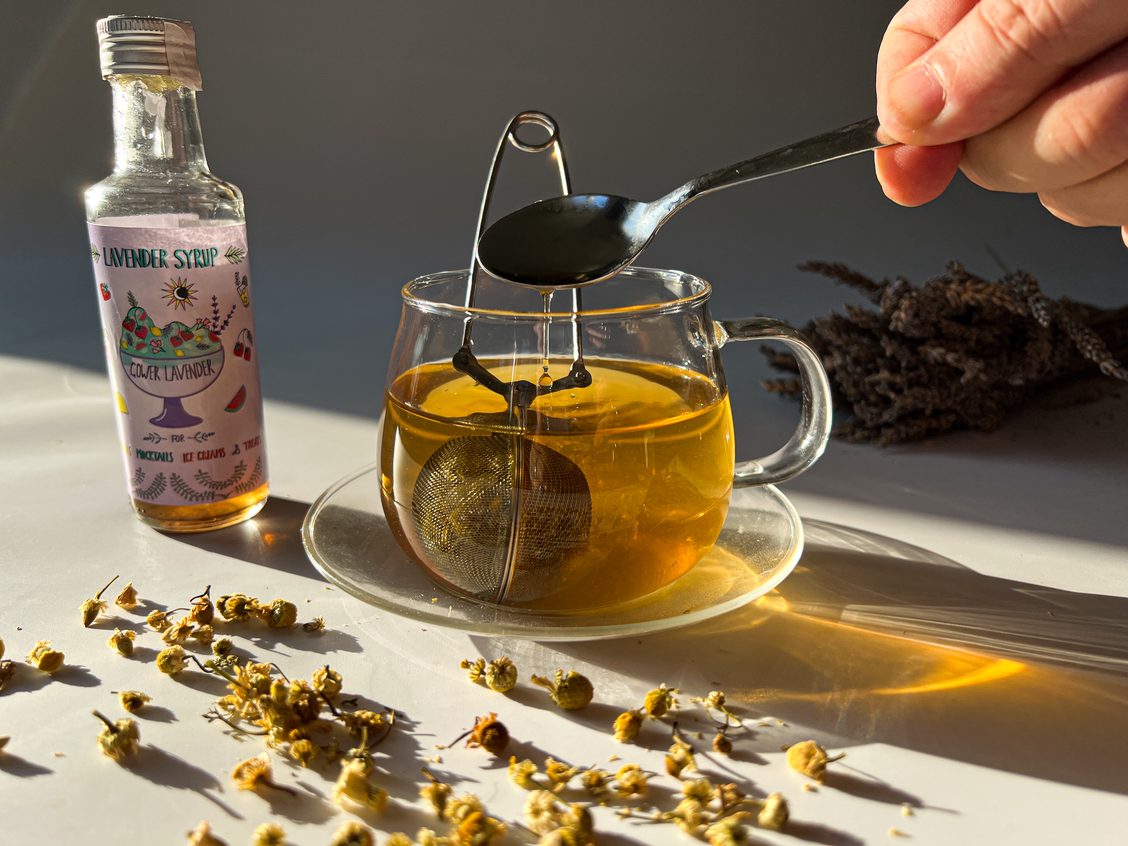 A glass mug with a chamomile tea infuser, on a table top with a bottle of Gower Lavender syrup and some dried chamomile flowers, with the sunshine making patterns on the table through the mug of tea