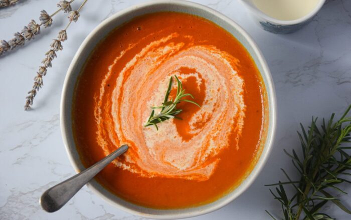 A white bowl filled with tomato, fennel and lavender soup, shown swirled with cream and with spoon resting in it