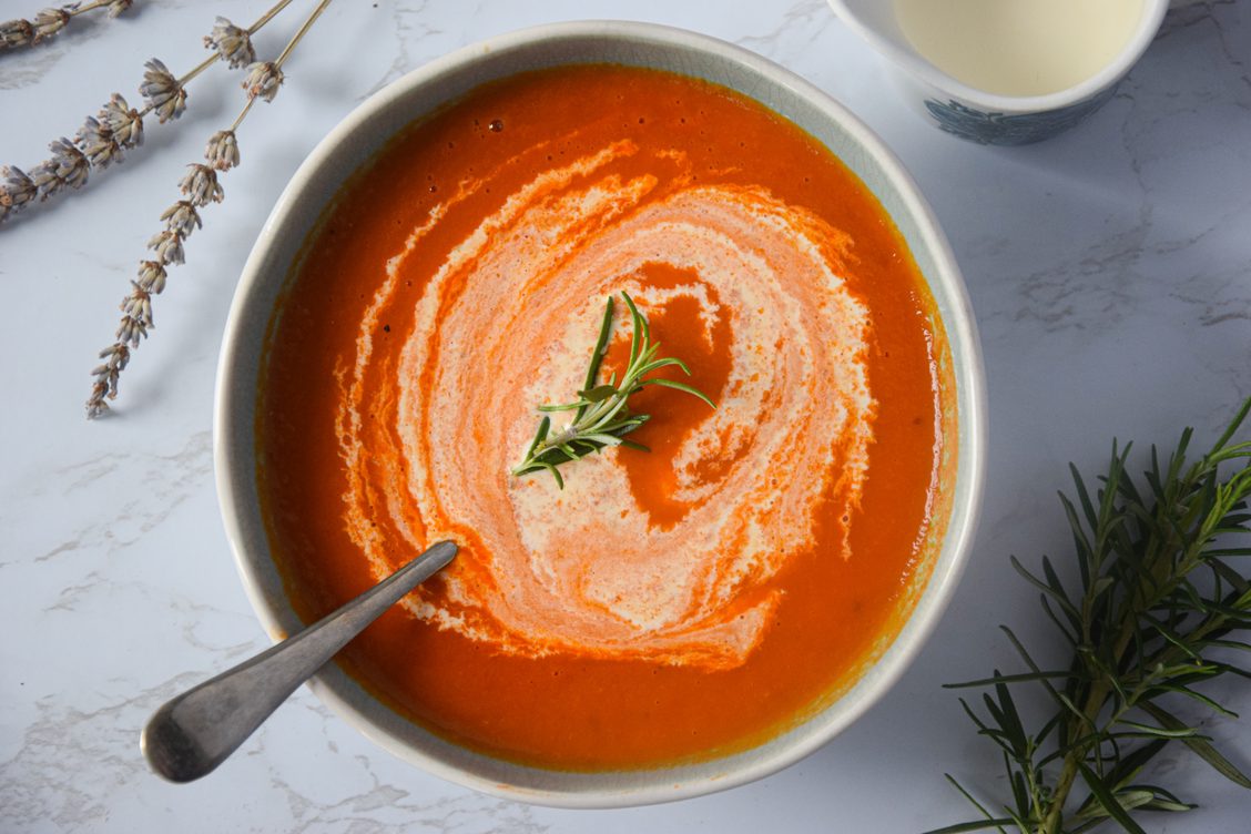 A white bowl filled with tomato, fennel and lavender soup, shown swirled with cream and with spoon resting in it