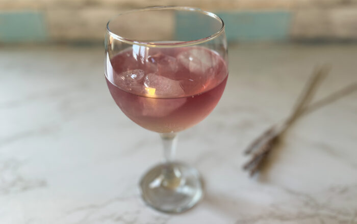 A pink cocktail in a stemmed glass with ice, on a marble table top, with dried lavender in the background