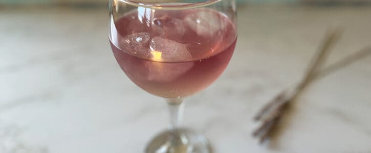 A pink cocktail in a stemmed glass with ice, on a marble table top, with dried lavender in the background