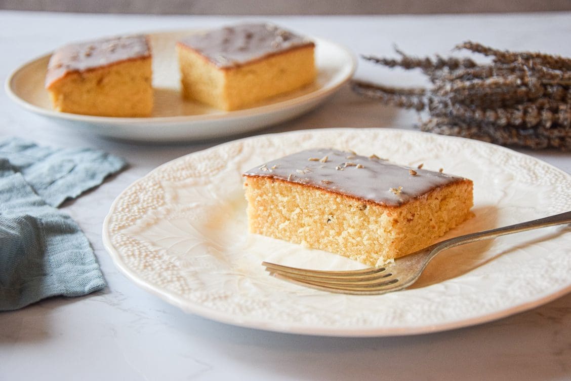A slice of lavender rose traybake cake served on a white plate with a silver fork