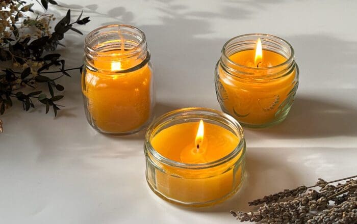 Three glass jars of different sizes containing beeswax lavender candles, each one lit, displayed on a white tabletop with a bunch of dried lavender