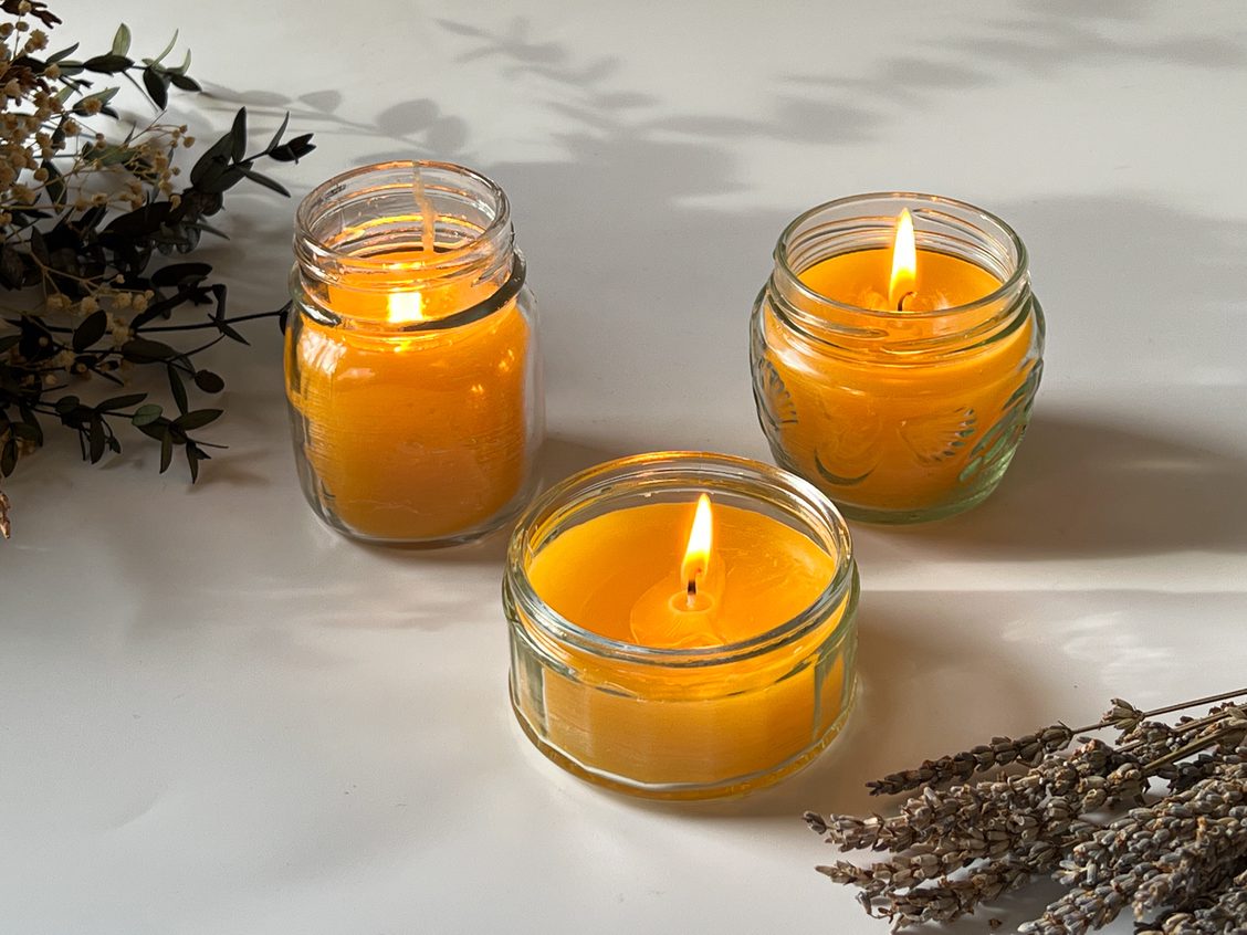 Three glass jars of different sizes containing beeswax lavender candles, each one lit, displayed on a white tabletop with a bunch of dried lavender