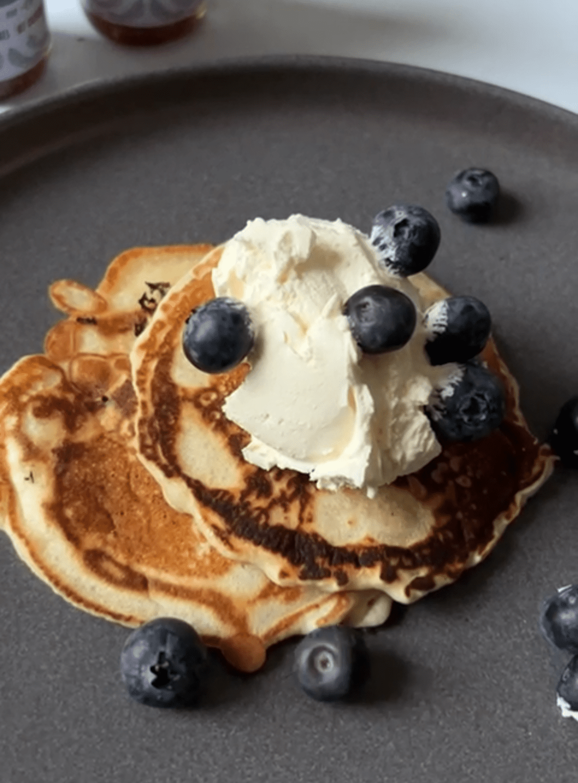 A stack of lavender pancakes on a dark grey plate, topped with sugared cream and blueberries
