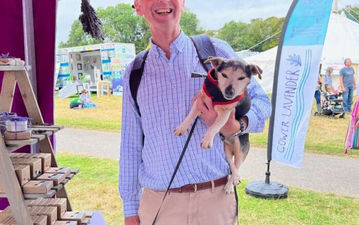 A smiling man in a blue shirt and red hat at the Gower Lavender stall, carrying a small dog under one arm