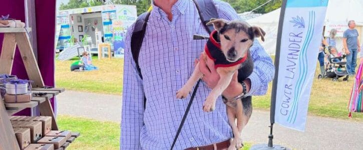A smiling man in a blue shirt and red hat at the Gower Lavender stall, carrying a small dog under one arm