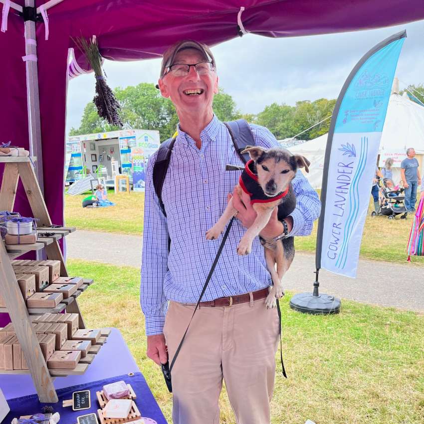 A smiling man in a blue shirt and red hat at the Gower Lavender stall, carrying a small dog under one arm